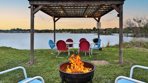 A wooden gazebo with a fire pit in the foreground, surrounded by outdoor seating and a view of a lake. - Waterfront RV LOT A (Willis)