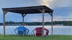A gazebo with a metal roof and wooden posts, surrounded by a grassy area with two chairs, one blue and one red, near a body of water. - Waterfront RV LOT A (Willis)