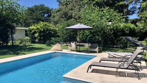 Una piscina rectangular con agua azul cristalina, rodeada por una solana de hormigón. Se ven sillas de descanso y una mesa con sombrilla al fondo.