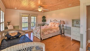 A bedroom with a wooden ceiling, a bed with floral bedding, a wicker chair, a small table with a lamp, and a white cabinet. - Serving Southern Charm at this Clarkesville House! (Clarkesville)