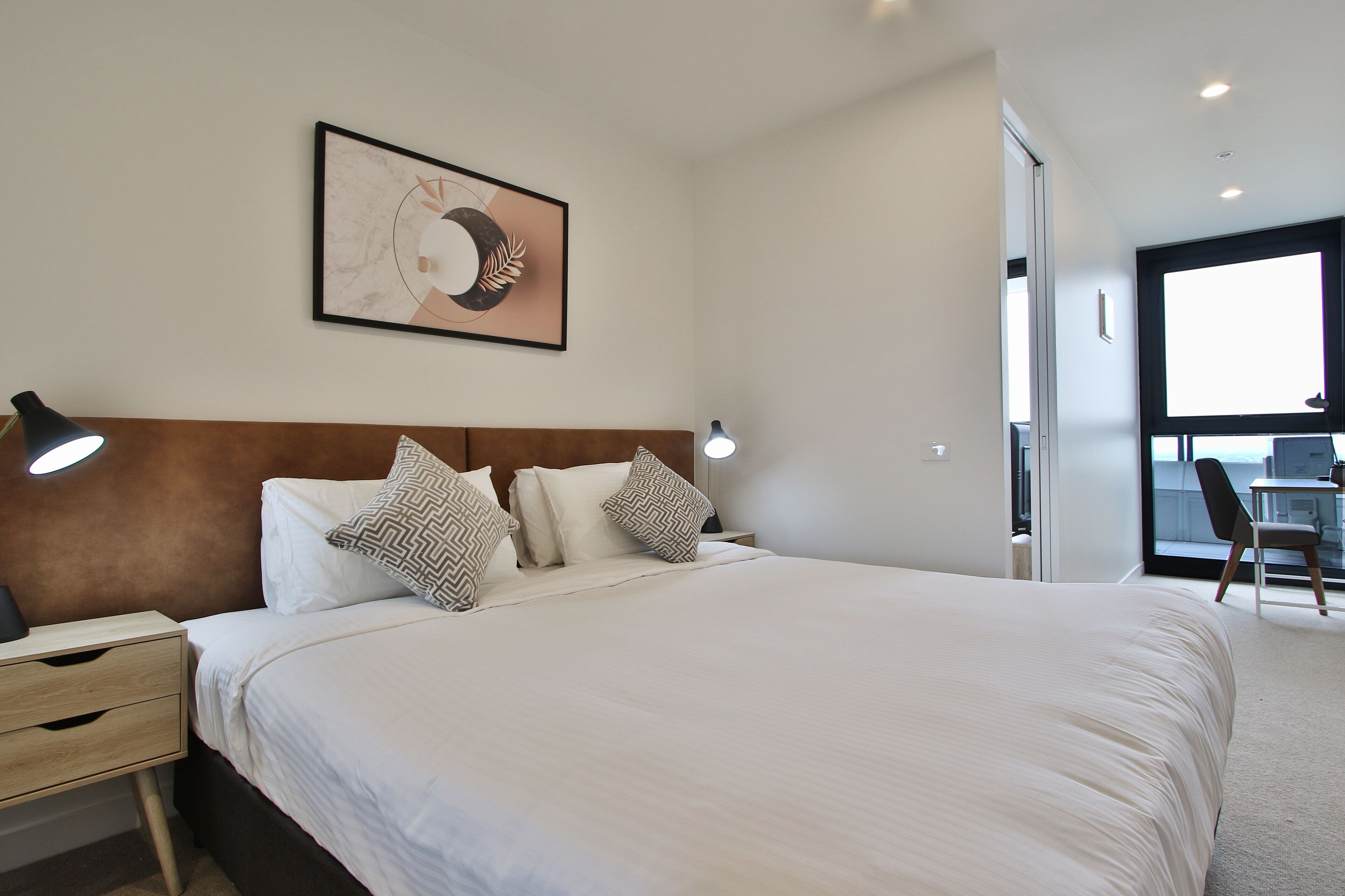A neatly made bed with white linens and patterned pillows, a headboard, bedside lamps, and a framed artwork on the wall.