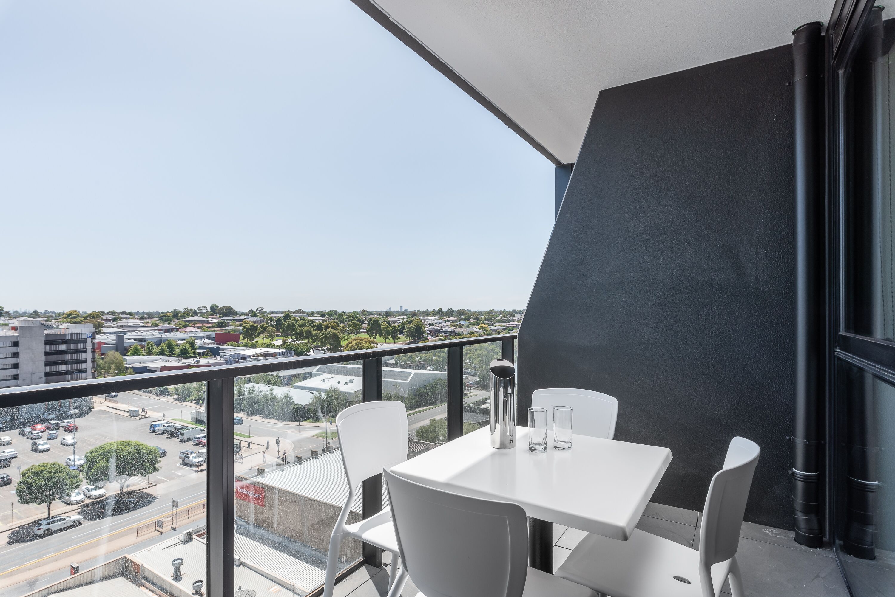 A balcony with a white table and chairs, a metal vase, and a clear view of a parking lot and buildings.