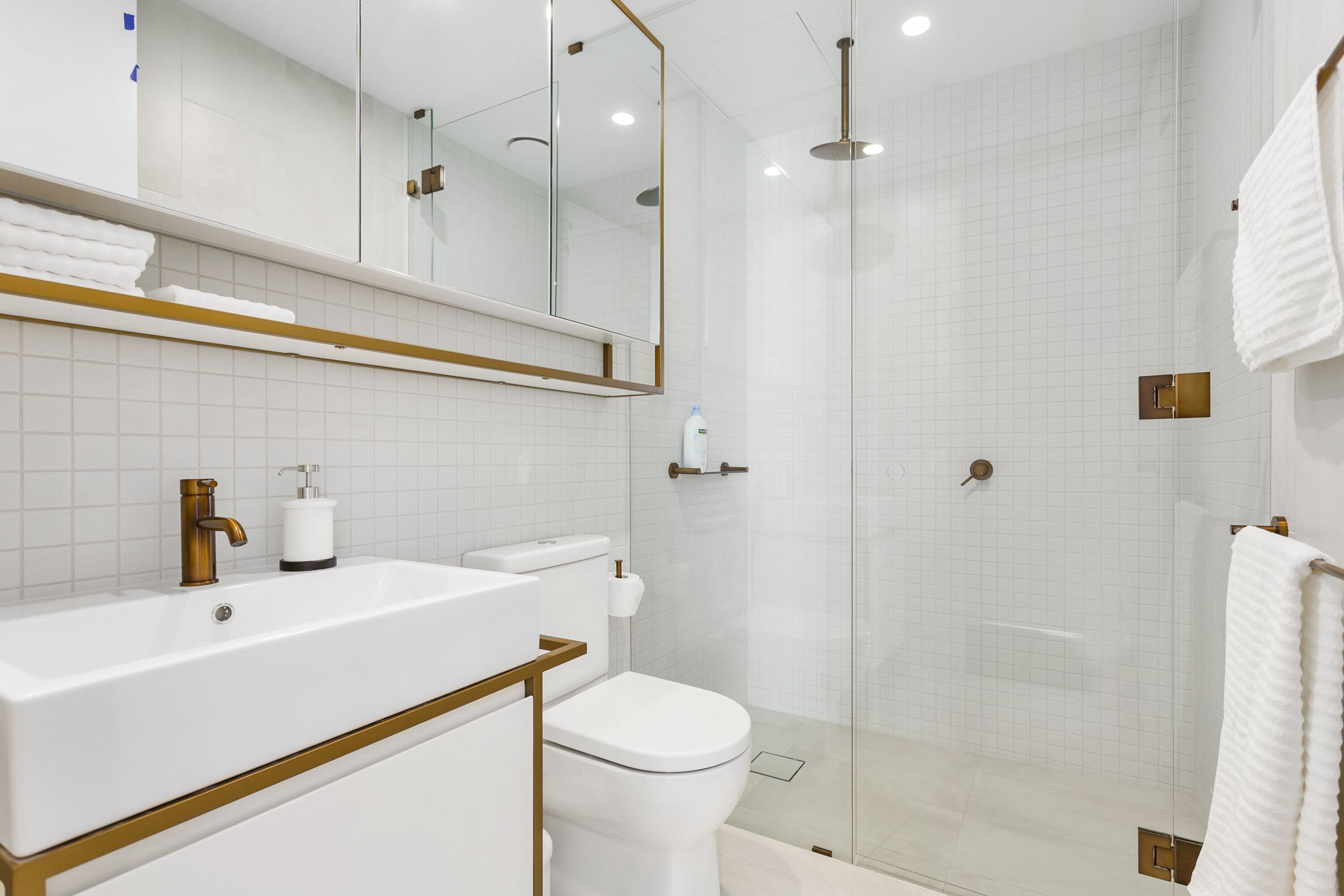 Un baño moderno con lavamanos blanco, detalles dorados, cabina de ducha con mampara de vidrio y un espejo grande.