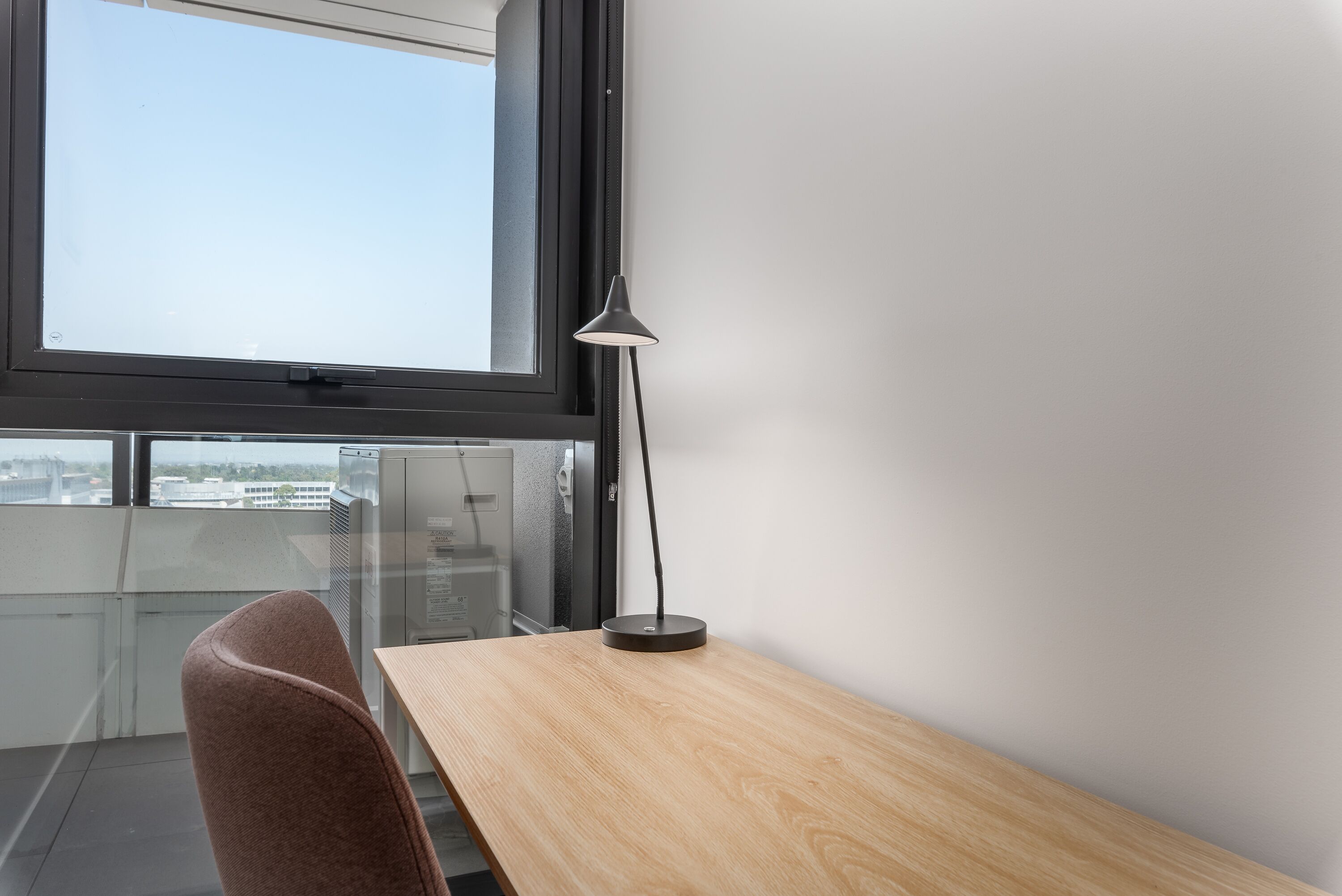 A modern office space with a wooden desk, a black desk lamp, and a clear view of the cityscape through a large window.