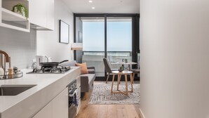 A modern kitchen with a gas stove, sink, and white cabinets. There is a dining area with a table and chairs, and a large window offering a view of the city. - Domi Serviced Apartments (Glen Waverley)