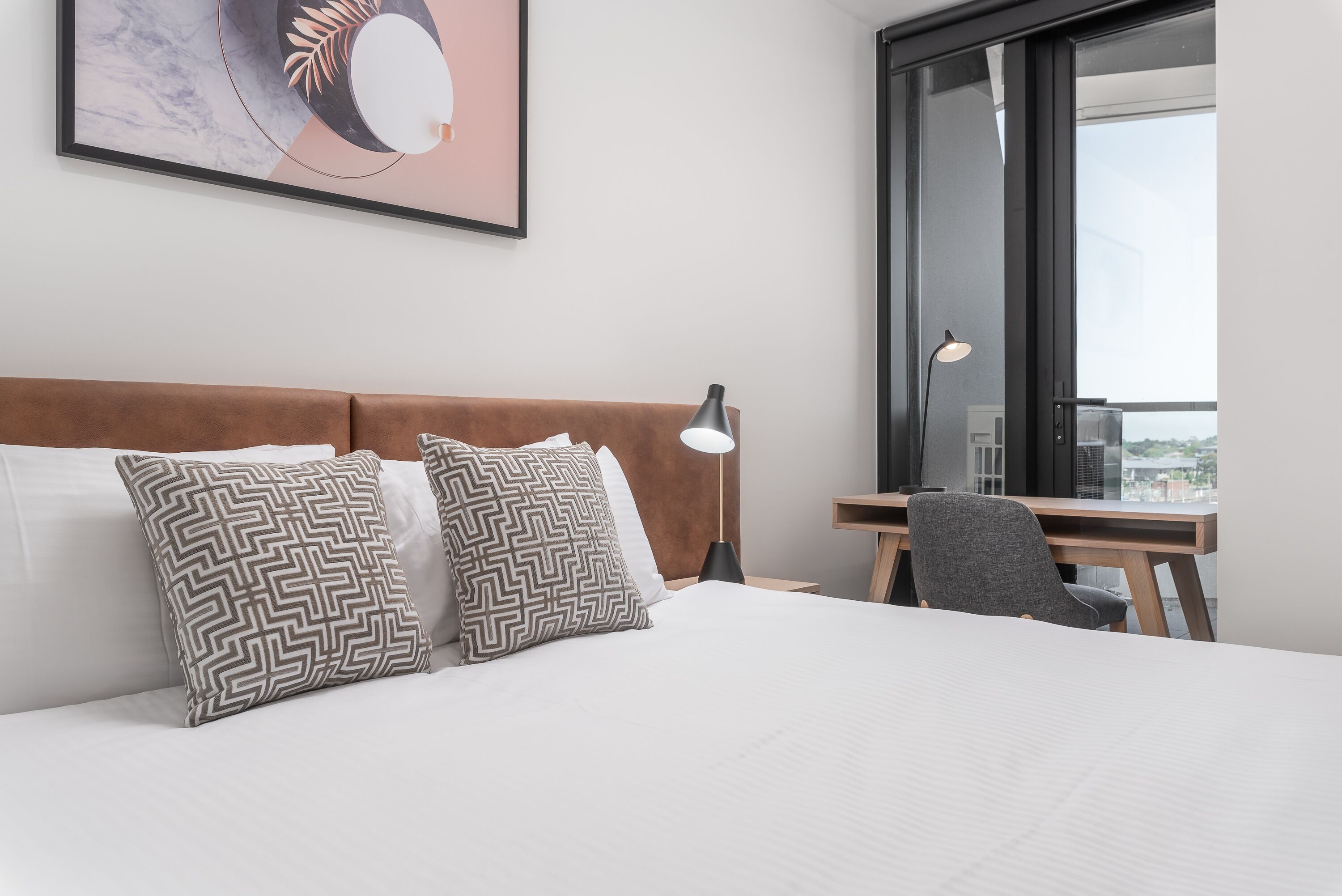 A hotel room with a bed, two patterned pillows, a desk with a chair, a lamp, and a large window with a view.