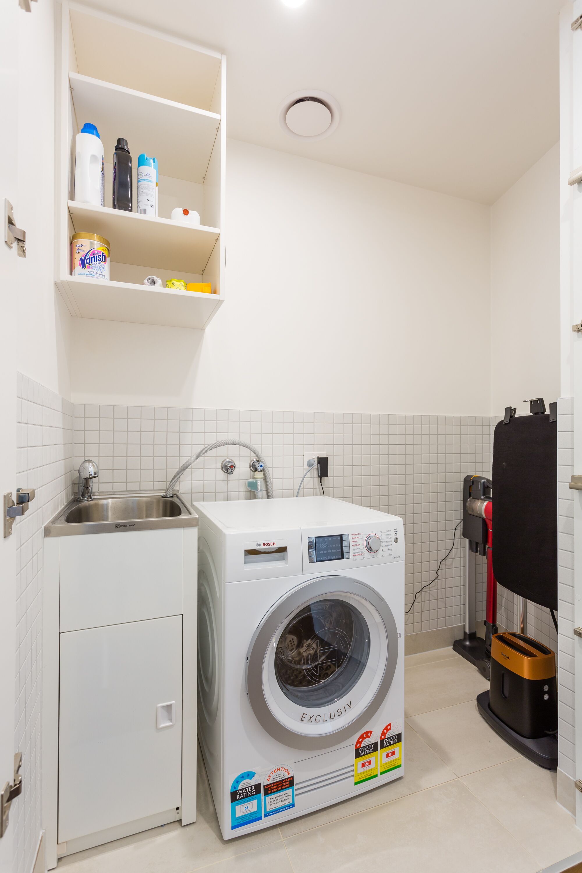 A compact laundry room with a Bosch washing machine, a sink, and a shelf with cleaning supplies.