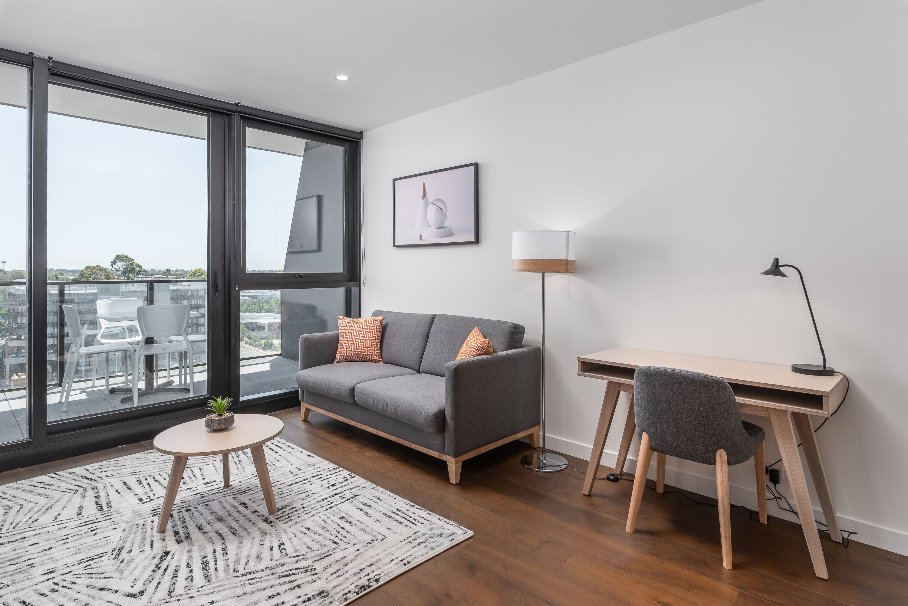 A modern living room with a grey sofa, a small round table, and a desk with a chair.
