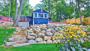 A blue cabin with a red flag, surrounded by a stone wall and a grassy area with yellow flowers. - SHORELINE CHATEAU-LAKEFRONT-LARGE DOCK-GAMEROOM- HOT TUB-WALK OUT DECK-LAKE VIEW (Fort Ann)