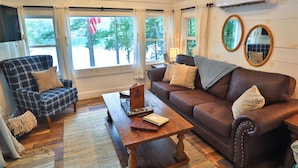 A cozy living room with a plaid armchair, a brown leather sofa, a wooden coffee table, and a large window with curtains. - SHORELINE CHATEAU-LAKEFRONT-LARGE DOCK-GAMEROOM- HOT TUB-WALK OUT DECK-LAKE VIEW (Fort Ann)