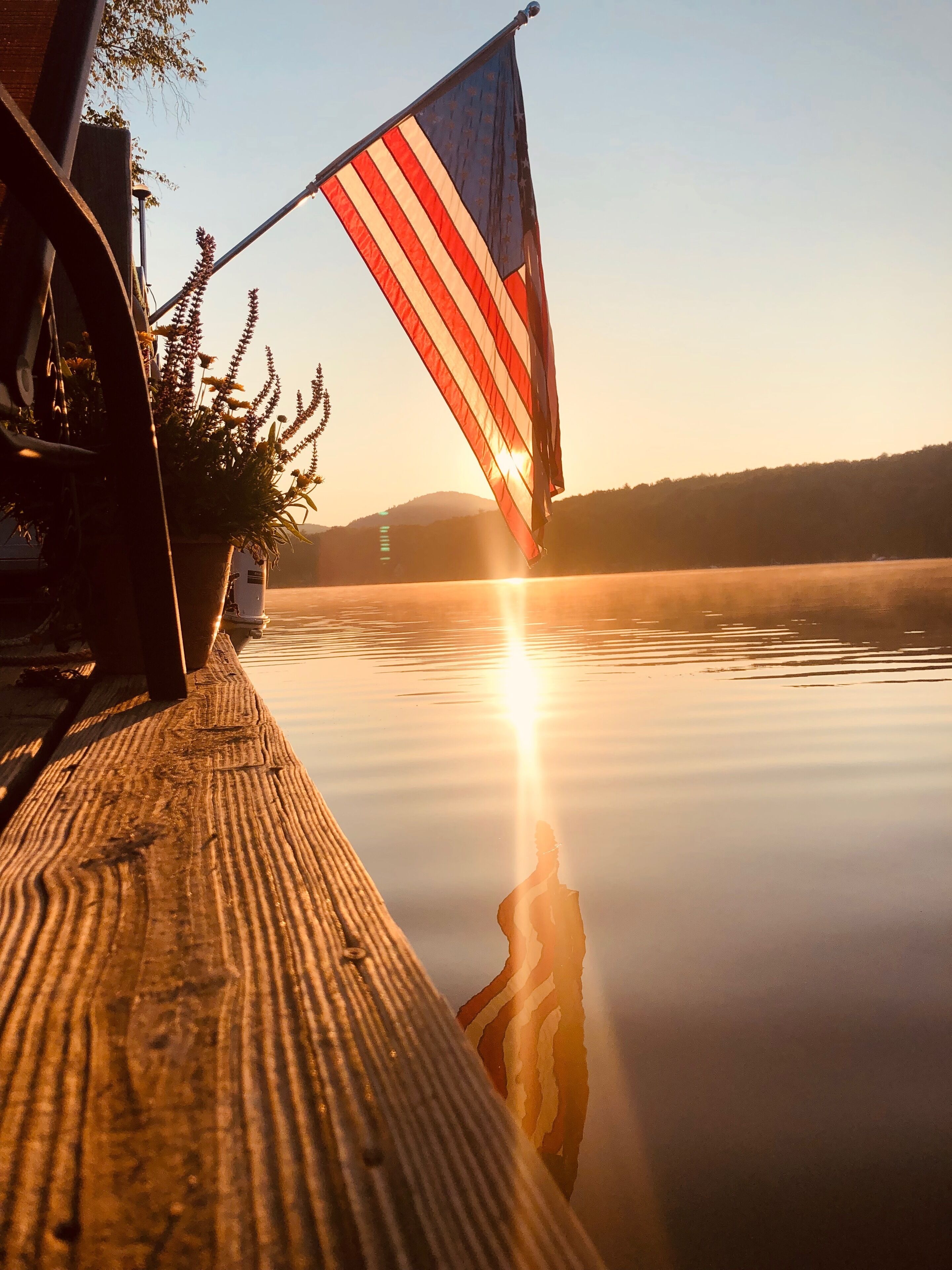 A wooden dock with a flag, calm water, and a sunset.