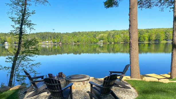 A lakeside outdoor seating area with Adirondack chairs and a circular table, surrounded by a stone fire pit. - SHORELINE CHATEAU-LAKEFRONT-LARGE DOCK-GAMEROOM- HOT TUB-WALK OUT DECK-LAKE VIEW (Fort Ann)
