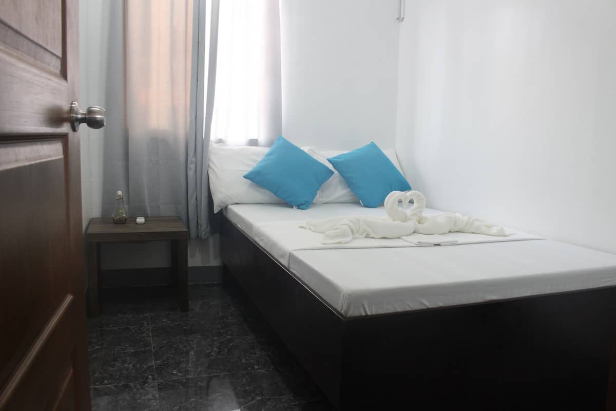 A small, neatly made bed with white bedding and two blue pillows, a wooden nightstand with a small lamp, and a door with a silver handle.