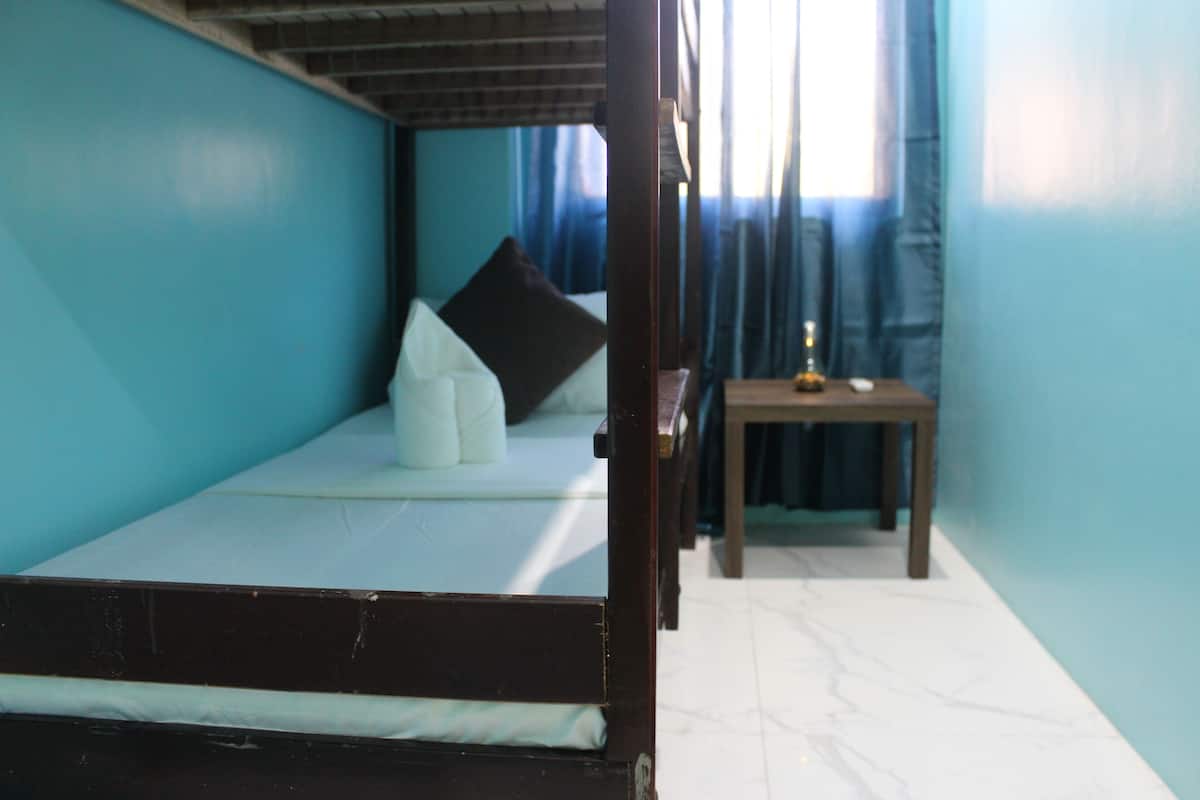 A single bed with a mattress and pillows, a wooden nightstand with a small table, and a window with curtains.