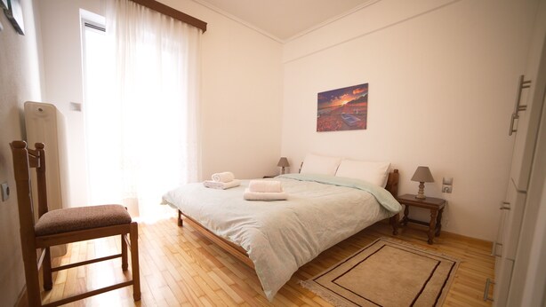 Een slaapkamer met houten vloer, een bed met witte lakens, een stoel, een nachtkastje met een lamp en een schilderij aan de muur.