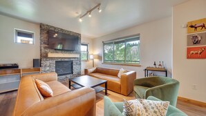 A modern living room with a stone fireplace, leather sofas, and a large window offering a view of greenery. - Four-Season Winter Park Condo w/ Pool Access! (Winter Park)