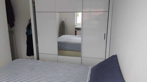 A bedroom with a bed, a wardrobe, and a mirror reflecting a window and a small table. - Apto 2 Quartos na Praia da Cachoeira (Florianópolis)
