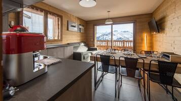 Eine moderne Küche mit einem roten Gerät, ein Essbereich mit Holztisch und -stühlen und Blick auf verschneite Berge durch große Fenster.