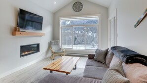 A modern living room with a fireplace, a large window with a snowy view, a flat-screen TV, and a cozy sofa. - New 2Bd 2Bth , Ski/Ski Out Condo with Private Hot Tub! (Sun Peaks)