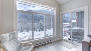 A room with a large window offering a view of a snowy landscape, a chair, and a heater under the window. - New 2Bd 2Bth , Ski/Ski Out Condo with Private Hot Tub! (Sun Peaks)