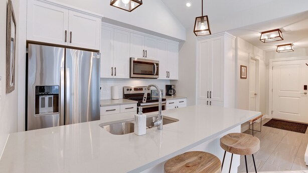 A modern kitchen with white cabinetry, stainless steel appliances, and a large island with stools. - New 2Bd 2Bth , Ski/Ski Out Condo with Private Hot Tub! (Sun Peaks)