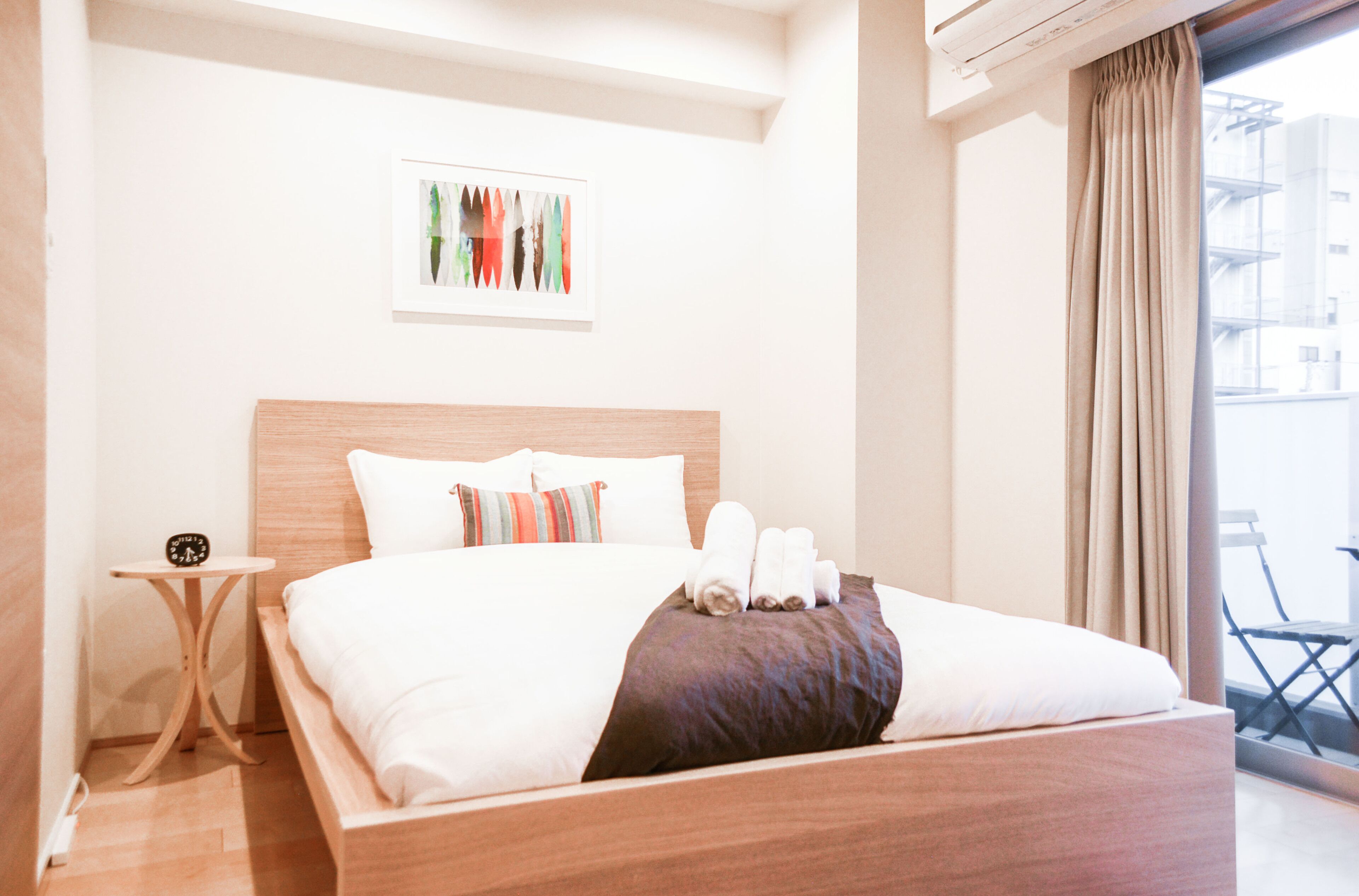 A hotel room with a wooden bed, white bedding, a small bedside table with a clock, a framed artwork on the wall, and a view of the city through a window with curtains.