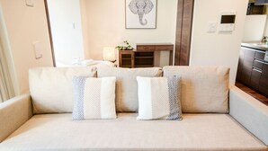 A living room with a beige sofa, two patterned throw pillows, a remote control on a tray, a wall-mounted light, and a framed elephant artwork.