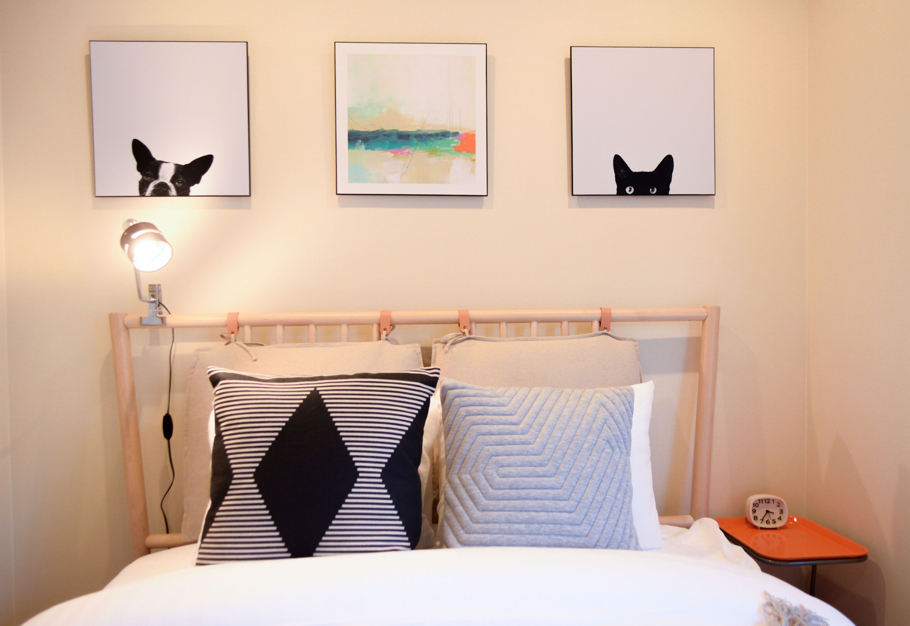 A neatly made bed with a striped pillow and a geometric pillow, a bedside lamp, and a small table with a clock.