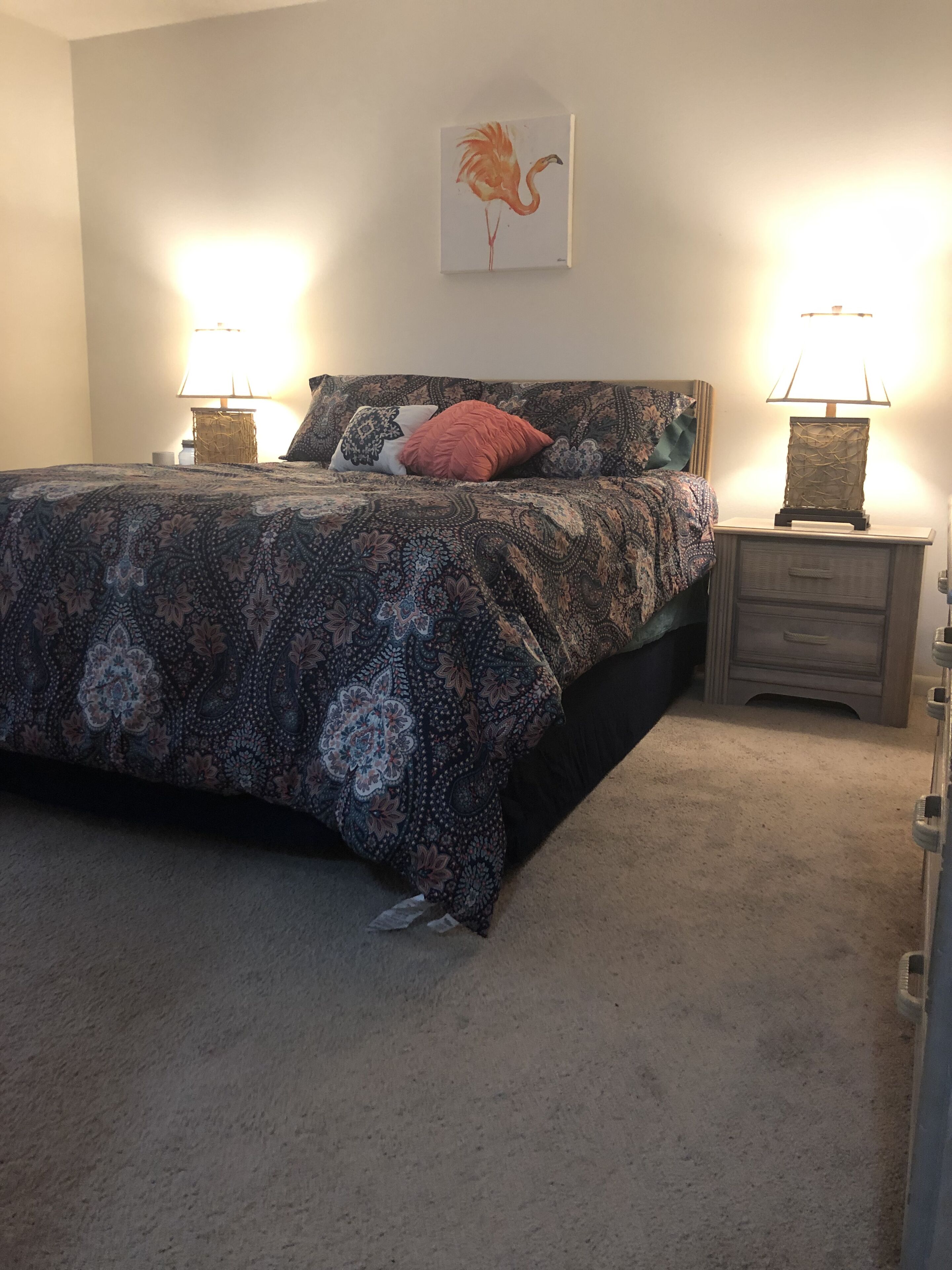 A bedroom with a patterned bedspread, two bedside lamps, a framed artwork on the wall, and a carpeted floor.