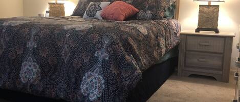 A bedroom with a patterned bedspread, two bedside lamps, a framed artwork on the wall, and a carpeted floor.