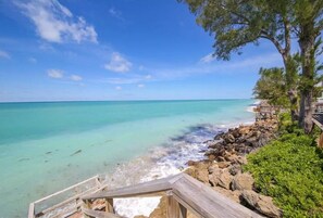 A wooden deck leading to a beach with clear turquoise water, rocky shoreline, and a tree-lined path.