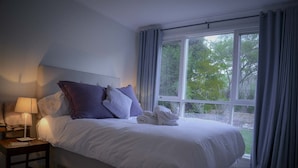 Een opgemaakt bed met witte lakens en meerdere kussens, een bedlampje en uitzicht op groen door een groot raam met gordijnen.