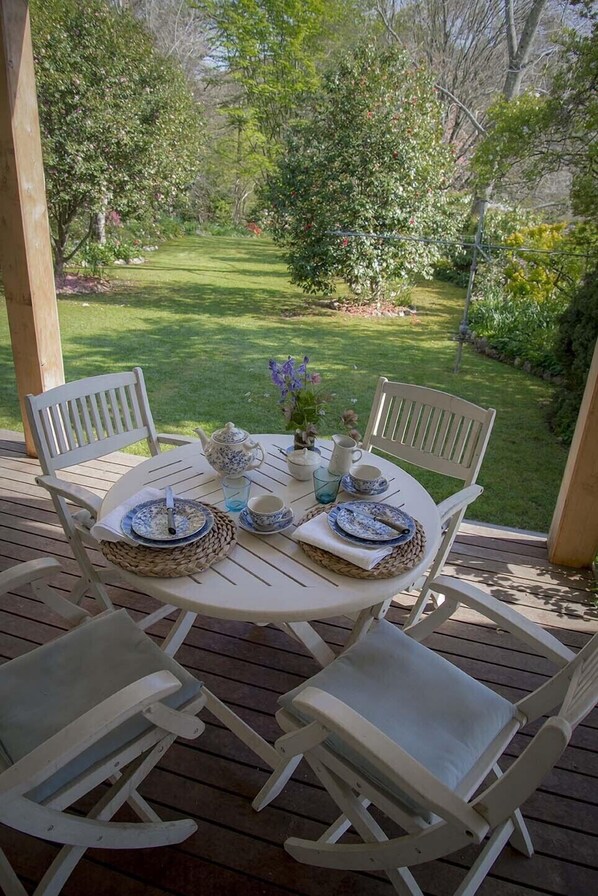 A round table with white chairs on a wooden deck, set for a meal with plates, cups, and a pitcher. - Mrs Simpson Cottage in Norala Garden (Bright)