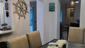 A dining area with a long table, beige upholstered chairs, a chandelier, a ship's wheel decoration, and a sign that reads "Memories made at the beach last a lifetime."