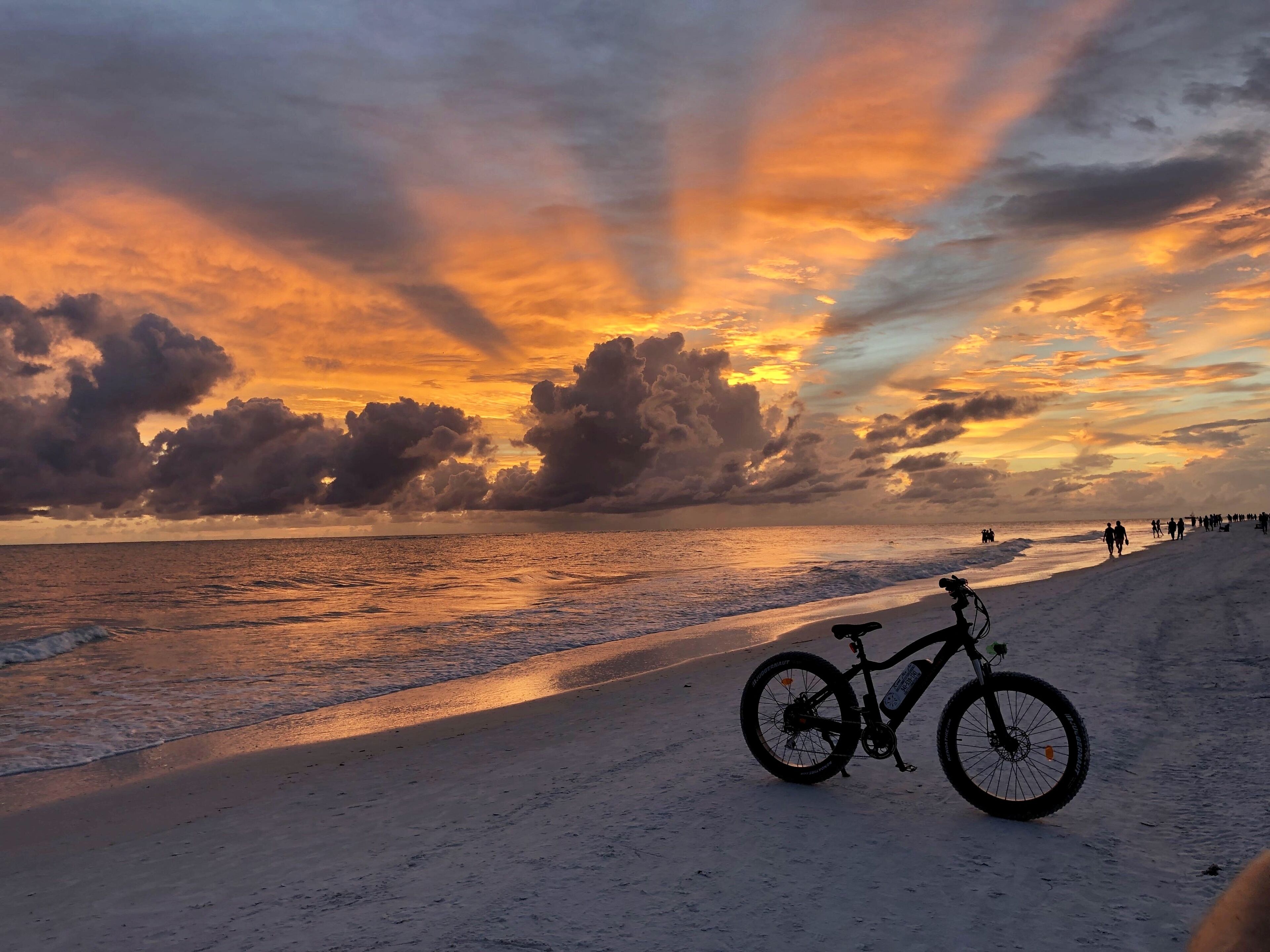 Una playa con una bicicleta estacionada en la arena, un cielo despejado y gente caminando a lo largo de la orilla.
