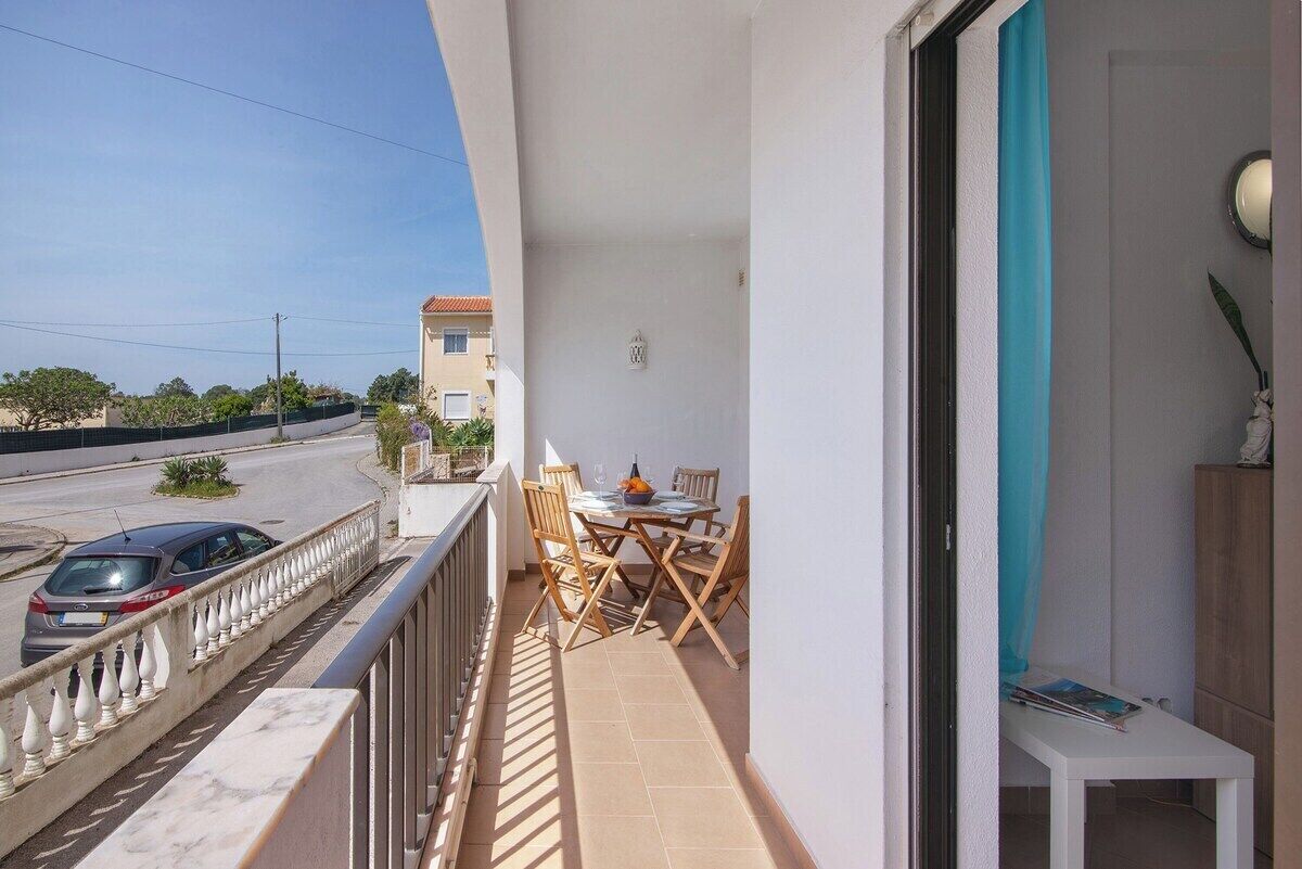 Varanda com mesa e cadeiras, uma mesinha com um abajur e vista para a rua.