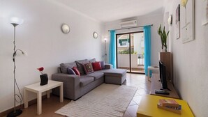 A living room with a grey sofa, a small white table, a yellow table, a floor lamp, and a TV.