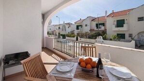 A balcony with a table set for two, a grill, and a view of white buildings with red roofs.