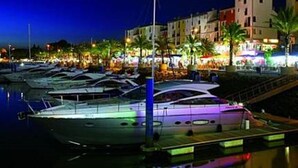 A marina at night with several boats docked, a waterfront promenade, and buildings with illuminated signs.