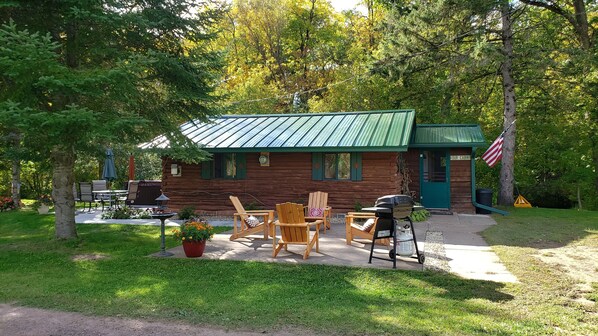 A rustic log cabin with a green metal roof, outdoor seating with chairs and a table, and a barbecue grill.