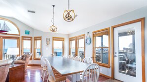 A spacious dining area with a long table and white chairs, large windows with wooden frames, and a view of the ocean. - Lightly Salted OBX, Beach front Luxury, Wild Horses, Pool, Hot Tub, 4x4 Beach (Corolla)