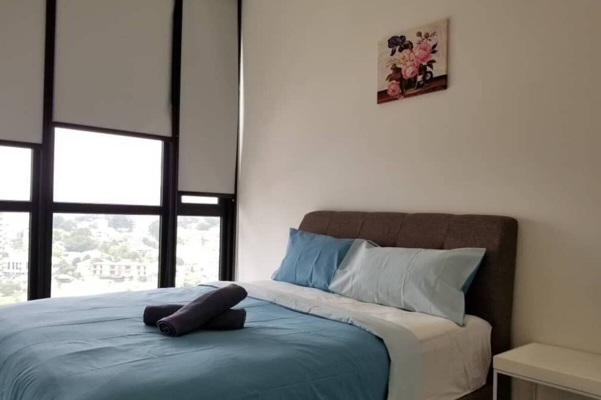 A bedroom with a large window, a bed with blue and white bedding, a brown headboard, and a small bedside table.