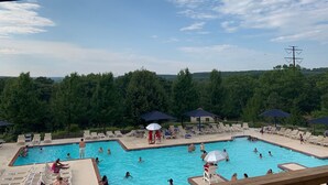 A swimming pool area with multiple pools, lounge chairs, umbrellas, and a forested background.