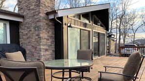 An outdoor deck with a glass-top table and four chairs, a stone wall, and a covered grill area.