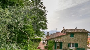 Una casa de piedra con techo de tejas, contraventanas verdes y un pequeño patio con un banco.