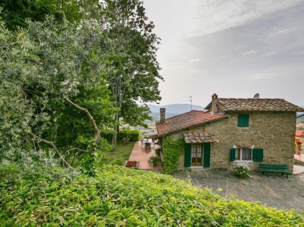 Ein Haus aus Stein mit Ziegeldach, grünen Fensterläden und einer kleinen Terrasse mit einer Bank.