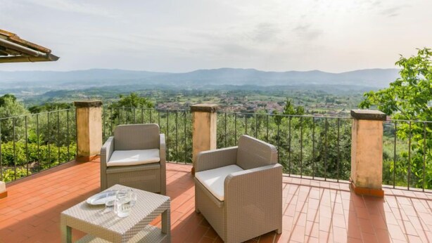 Una terraza con mobiliario de mimbre y una mesa, con vistas a un paisaje pintoresco con colinas y vegetación.