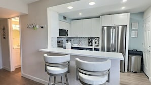 A modern kitchen with white cabinets, a stainless steel refrigerator, and a bar area with two stools. - Siesta Beach Condo Rental with direct beach and pool access (Siesta Key)