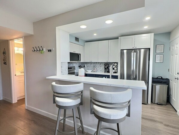 A modern kitchen with white cabinets, a stainless steel refrigerator, and a bar area with two stools. - Siesta Beach Condo Rental with direct beach and pool access (Siesta Key)