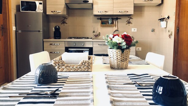 A modern kitchen with a microwave, stove, and cabinets. There is a dining table with striped placemats and a vase of flowers. - Residenza Donna Maria (Ercolano)